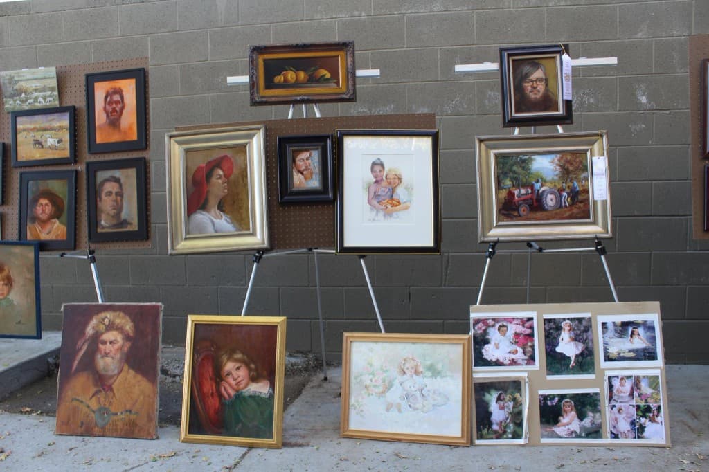 Framed oils and watercolors on tripods and pegboards against a cinderblock wall at an outdoor art setup
